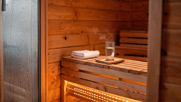 A warm cedar sauna interior viewed through the partially open glass door, a wooden bench with a neatly folded towel and a…