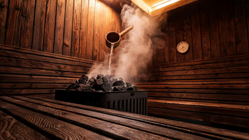 Interior of a traditional Finnish cedar sauna viewed from the lower bench, a wooden ladle mid-pour over a pile of dark sauna…