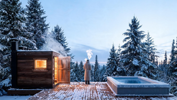 A person in a robe beside a steaming sauna and a sleek insulated cold plunge on a snow-dusted cedar deck in Canadian winter