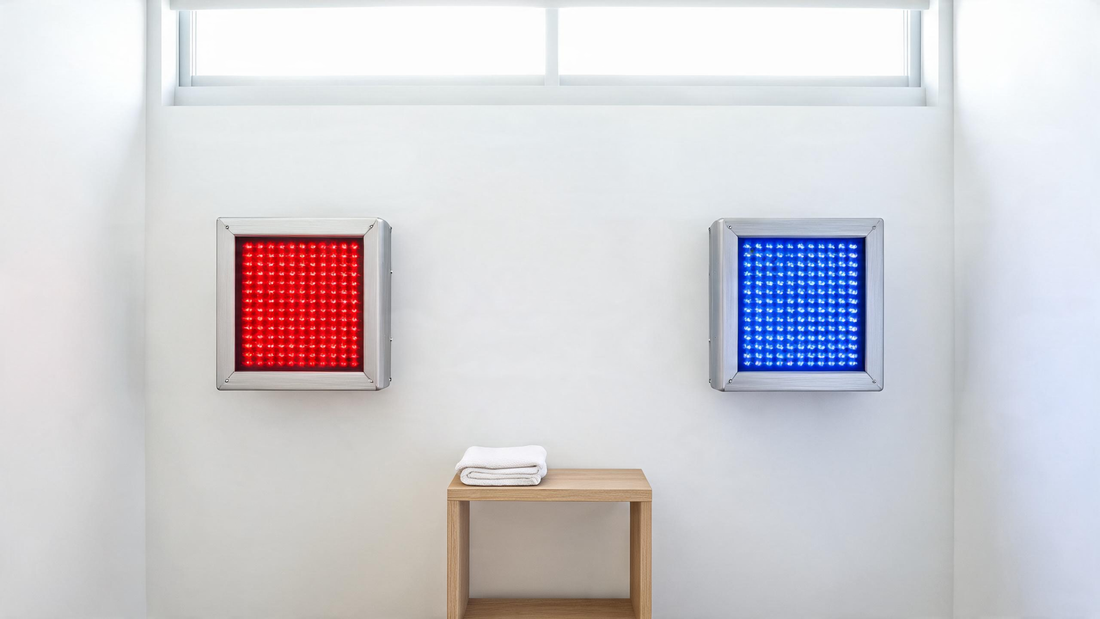 Two wall-mounted LED therapy panels side by side in a clean matte-white room, left panel glowing deep saturated red…