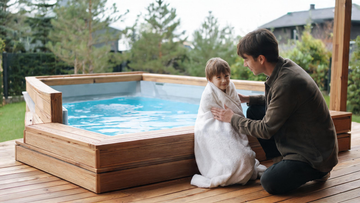 A parent kneeling beside a child wrapped in a warm towel near a small home cold plunge on a cedar deck, reassuring and calm, no submersion