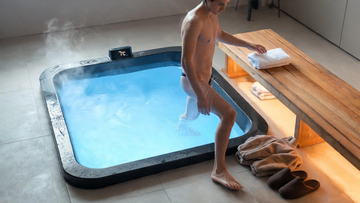 A person stepping calmly out of a sleek cold plunge in a tidy home wellness space, reaching for a folded towel on a warm cedar bench, faint vapour rising