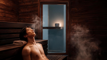 Person reclining eyes-closed on a cedar sauna bench, warm low light, faint steam, serene expression; a small speaker glowing softly outside the glass