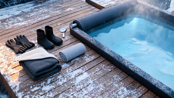 A flat-lay of cold plunge accessories on a snow-dusted cedar deck, neoprene gloves and booties, a fleece-lined robe, a floating thermometer, towel and insulated cover