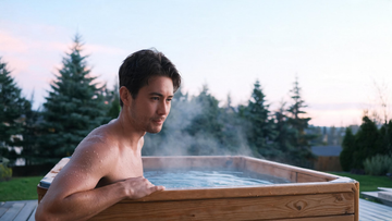 A composed adult easing slowly into a cedar-framed cold plunge in a calm Canadian setting, steam rising off the cold water, hand on the rim,