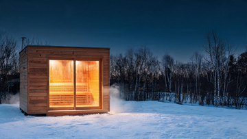 Hero, 16:9. A modern eco-friendly cedar sauna set in a snowy Canadian backyard at golden hour: warm amber interior glow…