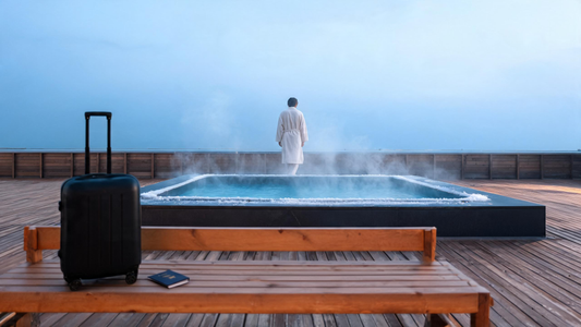 A traveller in a robe stepping toward a sleek insulated cold plunge on a cedar deck at first light, a wheeled suitcase and passport nearby