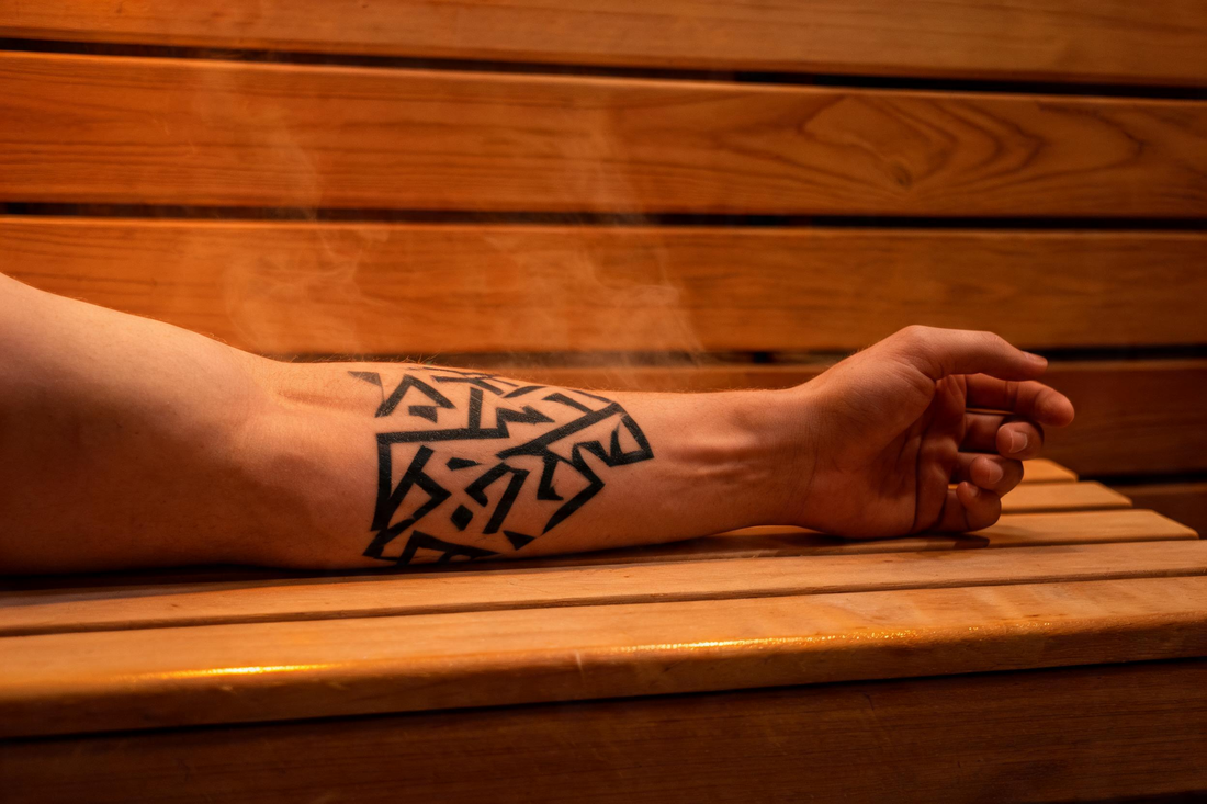 A close-up of a forearm with a bold geometric tattoo resting on the cedar bench of a warmly lit infrared sauna interior, the grain of the Grade-A Canadian