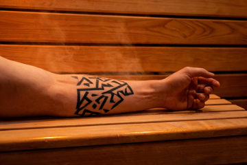 A close-up of a forearm with a bold geometric tattoo resting on the cedar bench of a warmly lit infrared sauna interior, the grain of the Grade-A Canadian