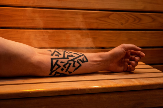 A close-up of a forearm with a bold geometric tattoo resting on the cedar bench of a warmly lit infrared sauna interior, the grain of the Grade-A Canadian