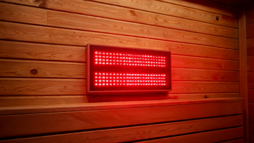 A Calore red-light therapy panel mounted on a cedar sauna wall, deep red and near-infrared LEDs glowing warmly across the panel face, warm cedar interior,