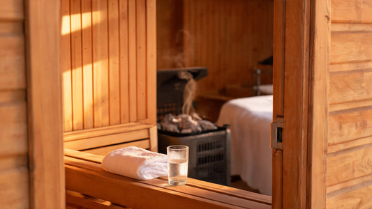 A warm cedar sauna interior viewed through the open door, a plush white towel folded on the bench and a glass of water resting on the lower bench, golden