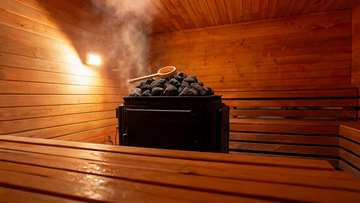 Interior of a cedar Finnish sauna, soft amber light on Grade-A Canadian cedar walls, a ladle resting on a Harvia heater piled with dark volcanic rocks, a