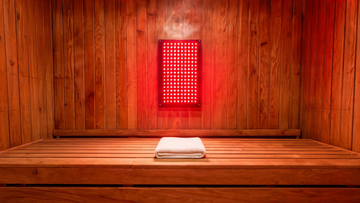 A cedar-lined sauna interior with a wall-mounted red light therapy panel glowing deep red against warm wood grain, a folded white towel on a cedar bench,