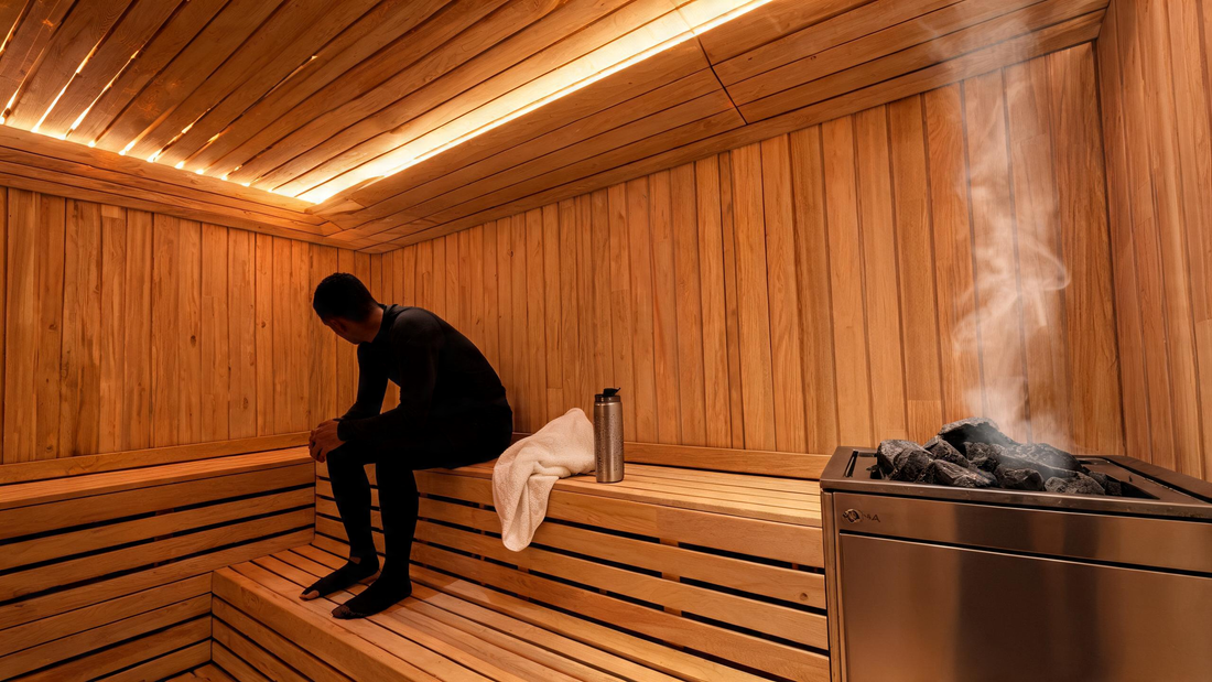 A person seated in a Grade-A Canadian cedar sauna interior, forearms resting on knees in post-workout relaxation posture, warm amber light from hidden LED