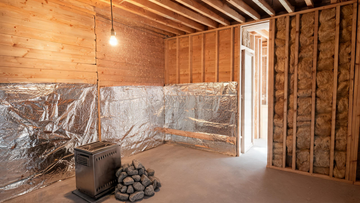 A partially built sauna room interior in mid-construction, cedar tongue-and-groove cladding half-installed on two walls over a foil-faced vapour barrier,