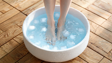 Bare feet being lowered into a white ceramic basin filled with clear water and ice cubes on a clean wooden deck,…