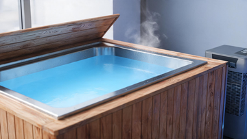 A sleek stainless-and-cedar cold plunge tub viewed at a low three-quarter angle, Glacier-blue water mirror-still with faint…