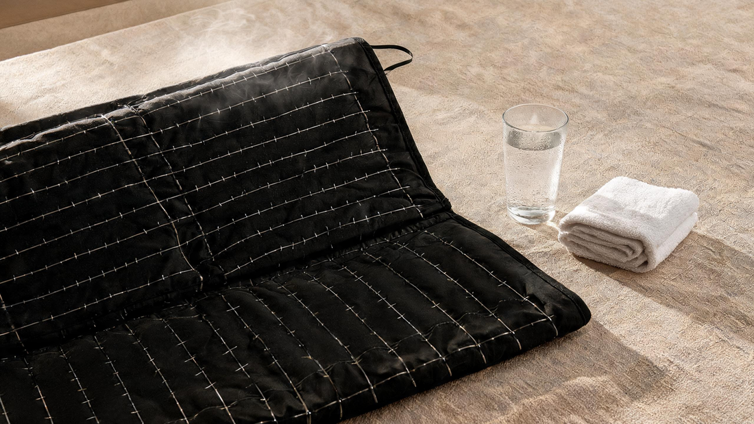 A rolled infrared sauna blanket lying open on a neutral linen surface, its dark interior and thin heating-wire texture…
