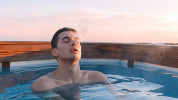 A calm person submerged to the shoulders in a sleek cold plunge at dawn, eyes closed, shoulders visibly softened (not braced), faint vapour